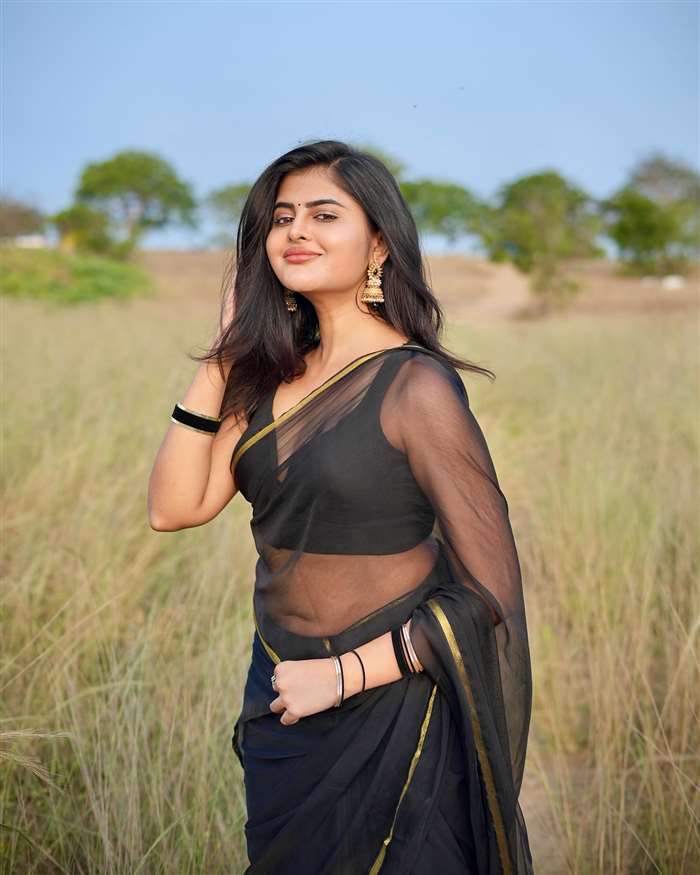 Rheena Krishnan is captured in a bright, backlit portrait leaning against a dark vertical structure, gazing directly at the camera with a gentle expression.