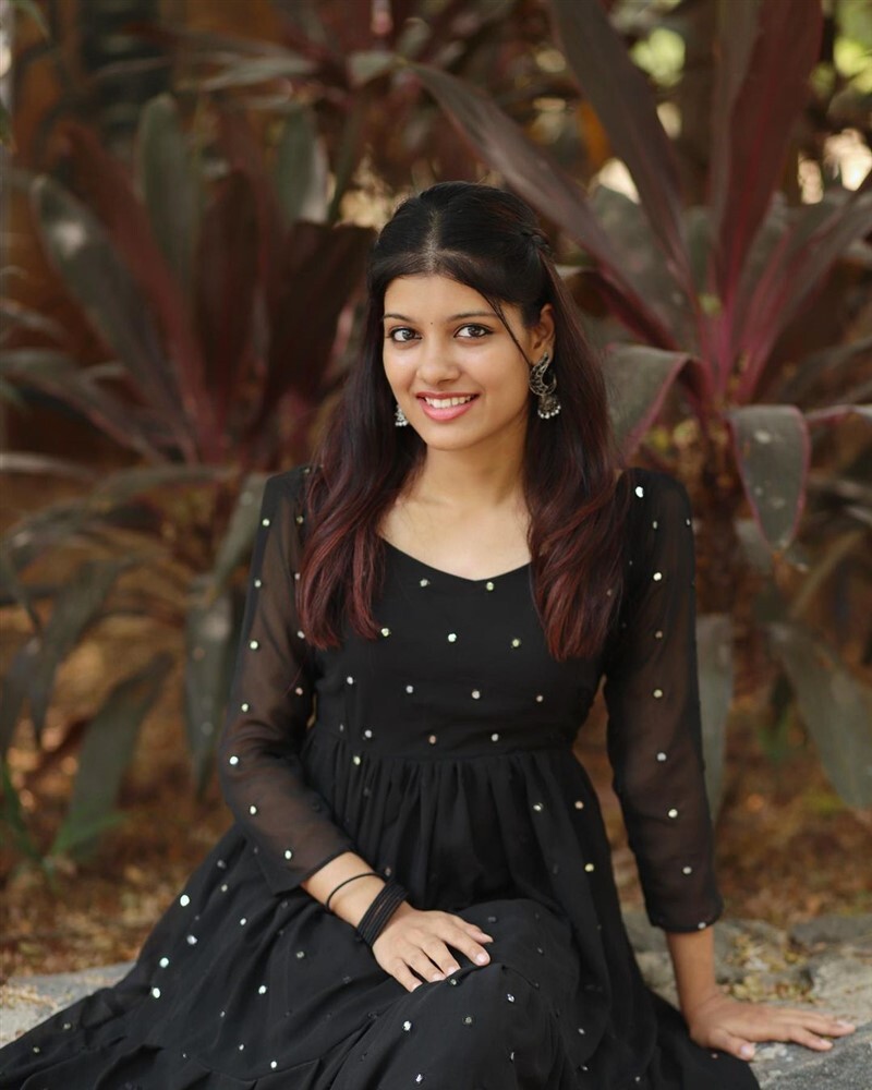 Monisha Blessy standing outdoors holding a large, open patterned umbrella and smiling gently.