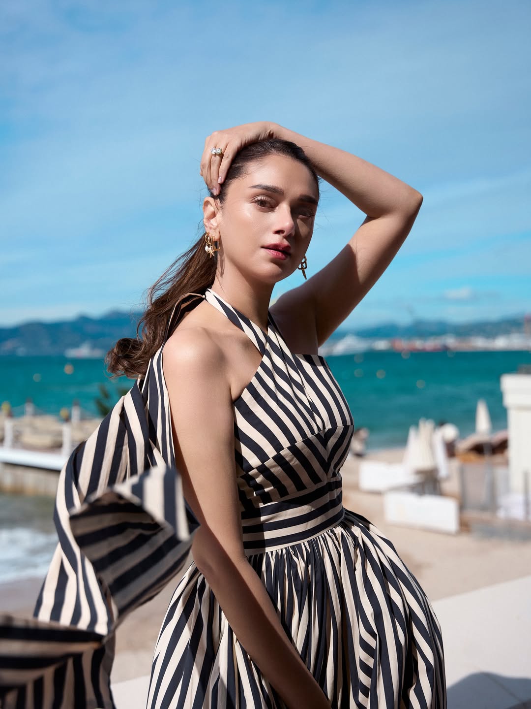 Aditi Rao Hydari standing on a railing overlooking the ocean with black and white striped attire and flowing dark hair.