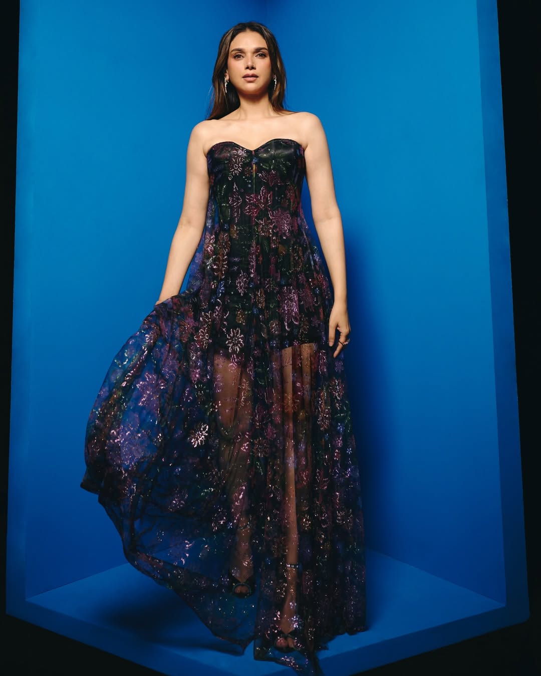 Aditi Rao Hydari sitting on a striped sofa wearing a flowing, richly patterned gown with deep purple and maroon tones.