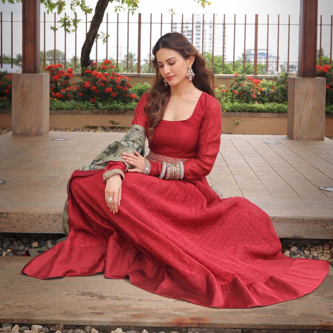 Amyra Dastur reclining on a wooden structure in a flowing red gown with green and gold accents.