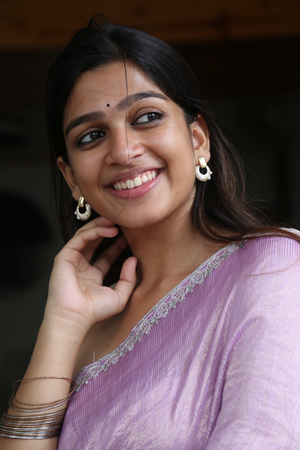 Ananthika Sanilkumar is leaning against a large brown tree, smiling outdoors while draped in a light top and purple fabric.