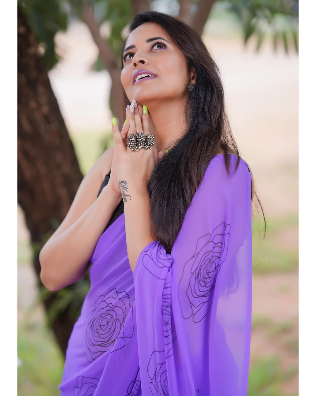 Anasuya Bharadwaj stands outdoors draped in a vibrant purple fabric with dark floral patterns and dark clothing.