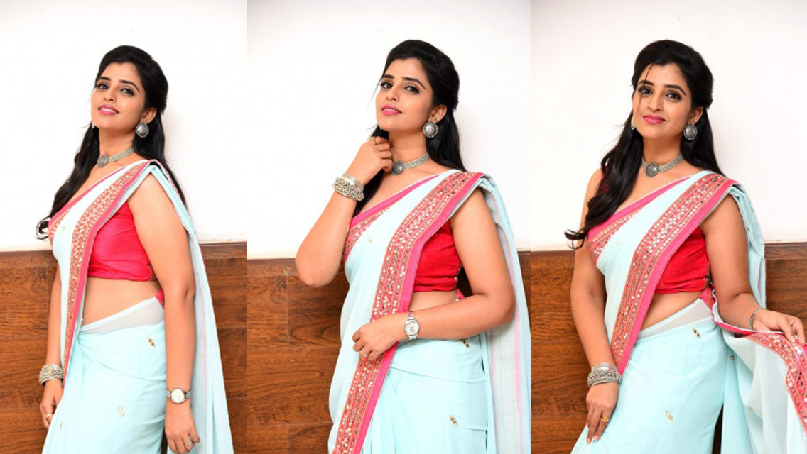 Syamala wearing a light blue kurta with a pink dupatta and red garment, leaning against a dark brown wooden surface.