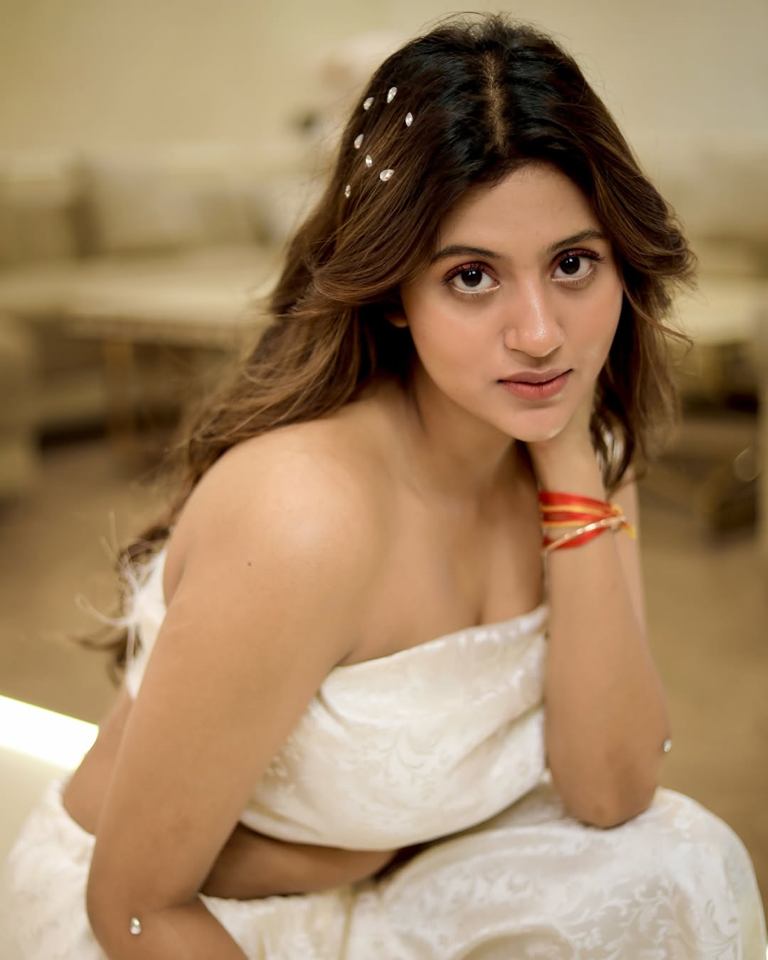 Anjali Arora standing indoors in a flowing light-colored gown with wavy brown hair and metallic sandals.