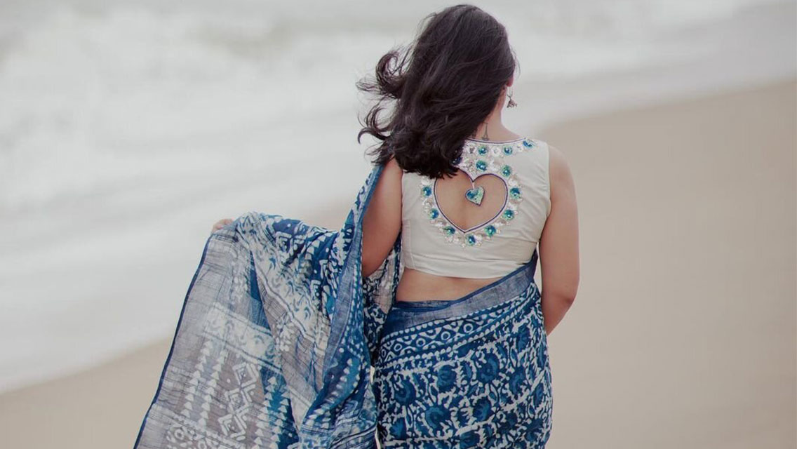 Anu Shivv stands barefoot on wet sand at the shoreline, draped in a flowing blue and white patterned garment.