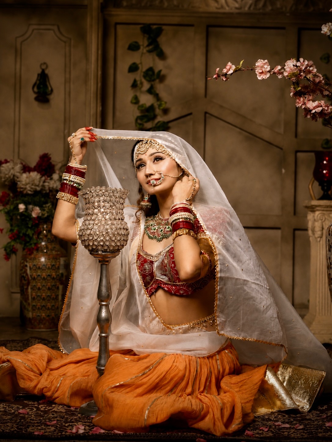 Arushi Nishank is captured in a profile view wearing elaborate gold jewelry and rich red attire against an ornate background.