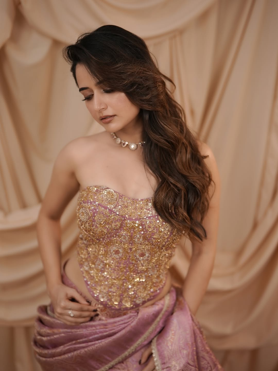 Ashika Ranganath stands in a dimly lit interior wearing a flowing purple gown against warm draped fabric.