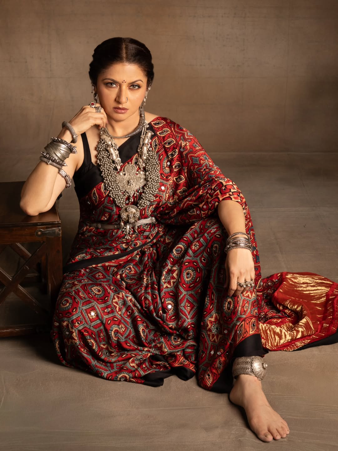 Bhagyashree is seated on the floor, draped in richly patterned fabric, posing in a studio portrait.