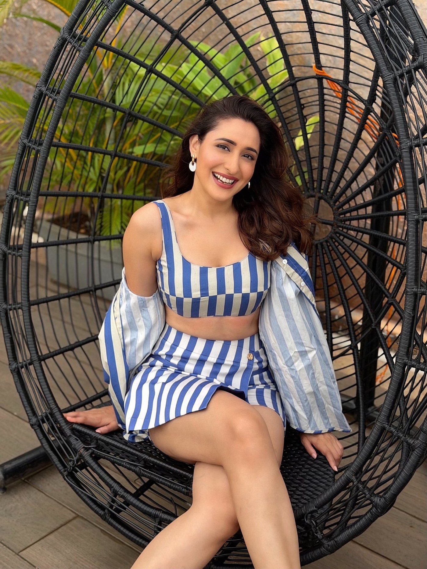 Pragya Jaiswal is captured seated outdoors against a dark metal structure, showcasing a beautiful striped two-piece ensemble. - Photos & Updates
