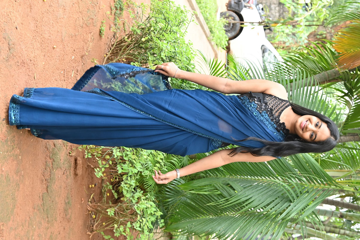 Brigida posing against a warm background in a flowing green saree with gold jewelry.