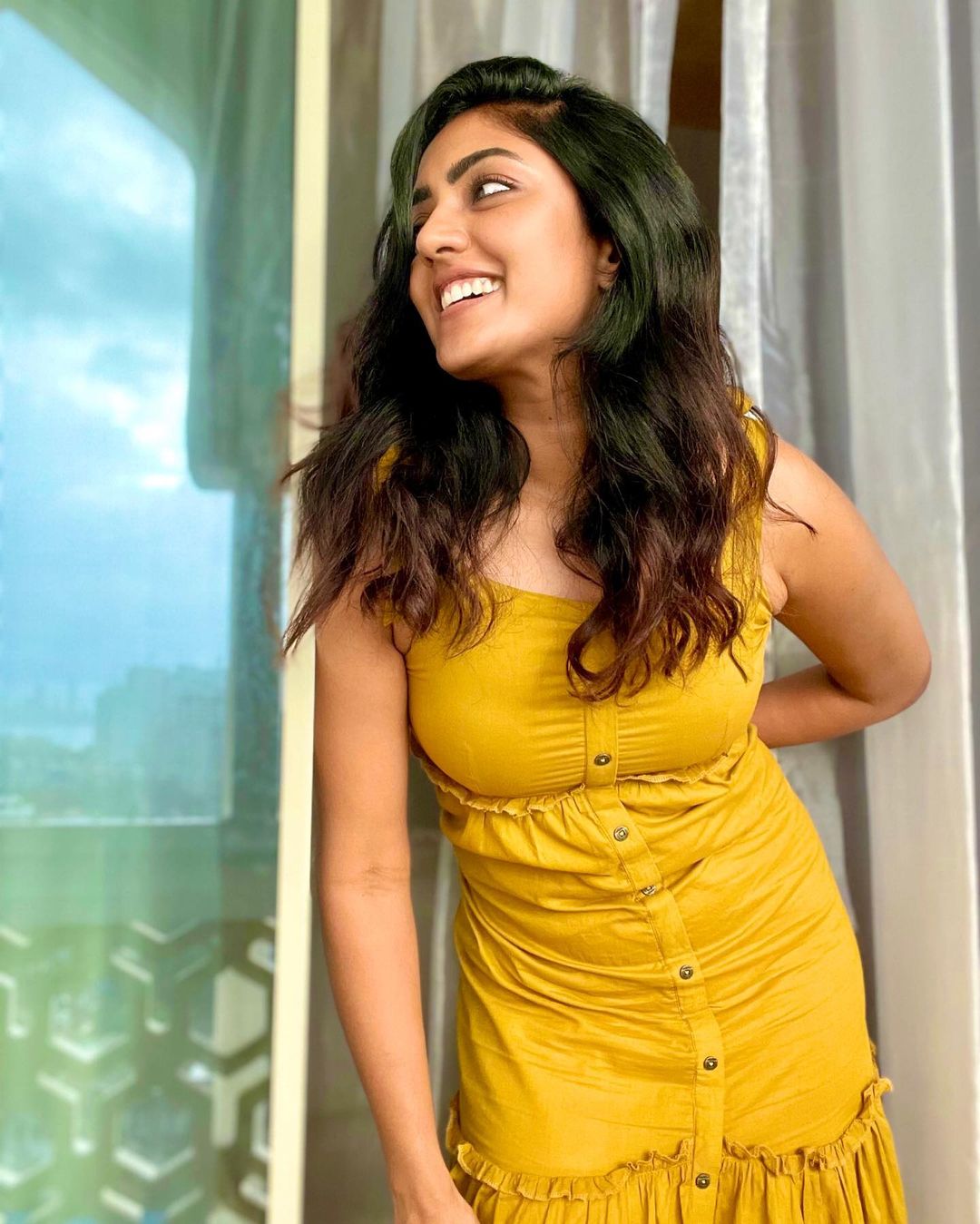Eesha Rebba in a bright yellow, textured sleeveless garment, smiling warmly indoors near a window.