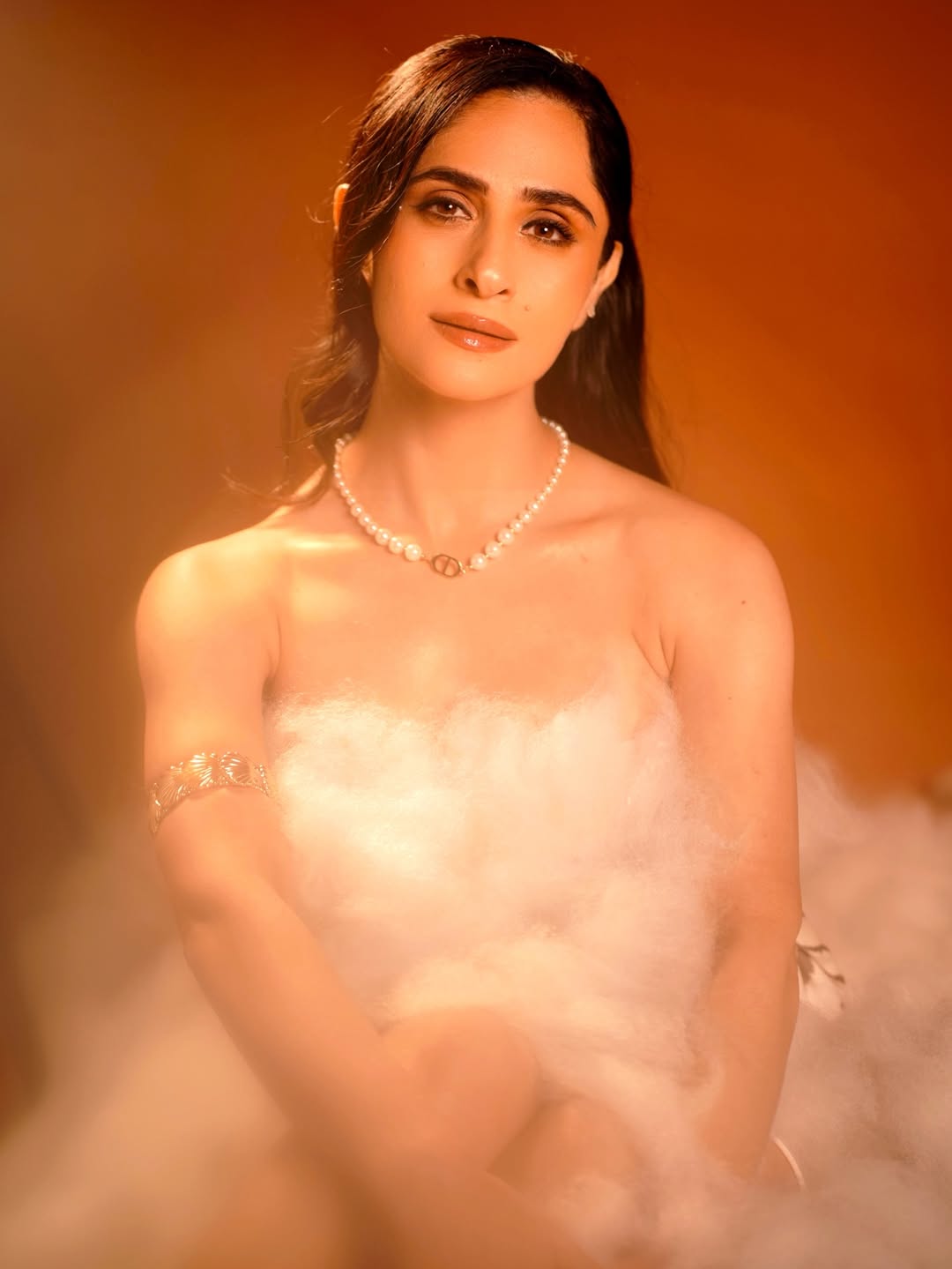 Hunar Hali is draped entirely in voluminous, soft white or off-white material, wearing delicate jewelry and reclining against a warm background.