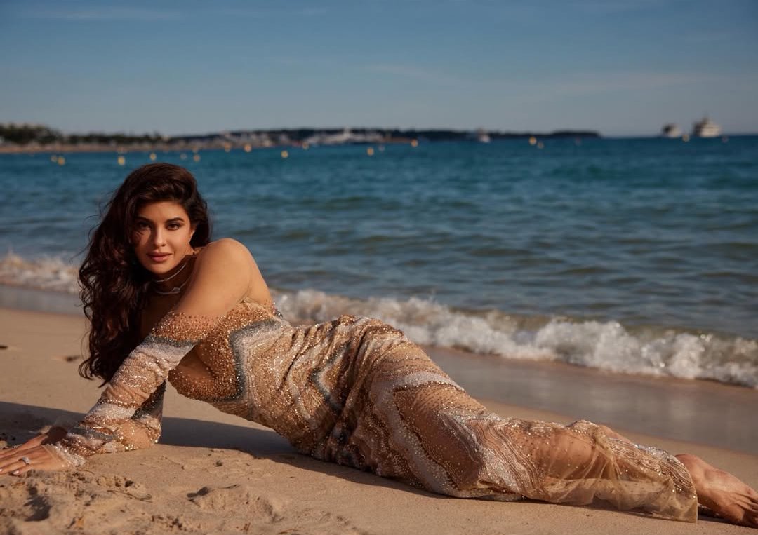 Jacqueline Fernandez stands on a wet sandy beach, partially submerged by gentle waves, wearing a shimmering gold gown.