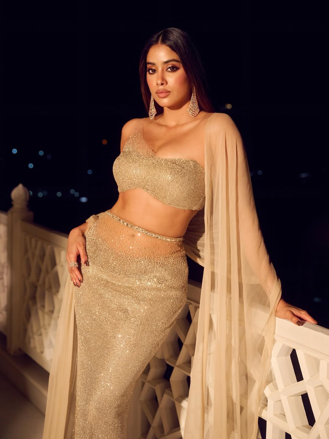 Janhvi Kapoor is sitting in a two-piece outfit of light gray/beige and vibrant orange, with long, flowing wavy hair.