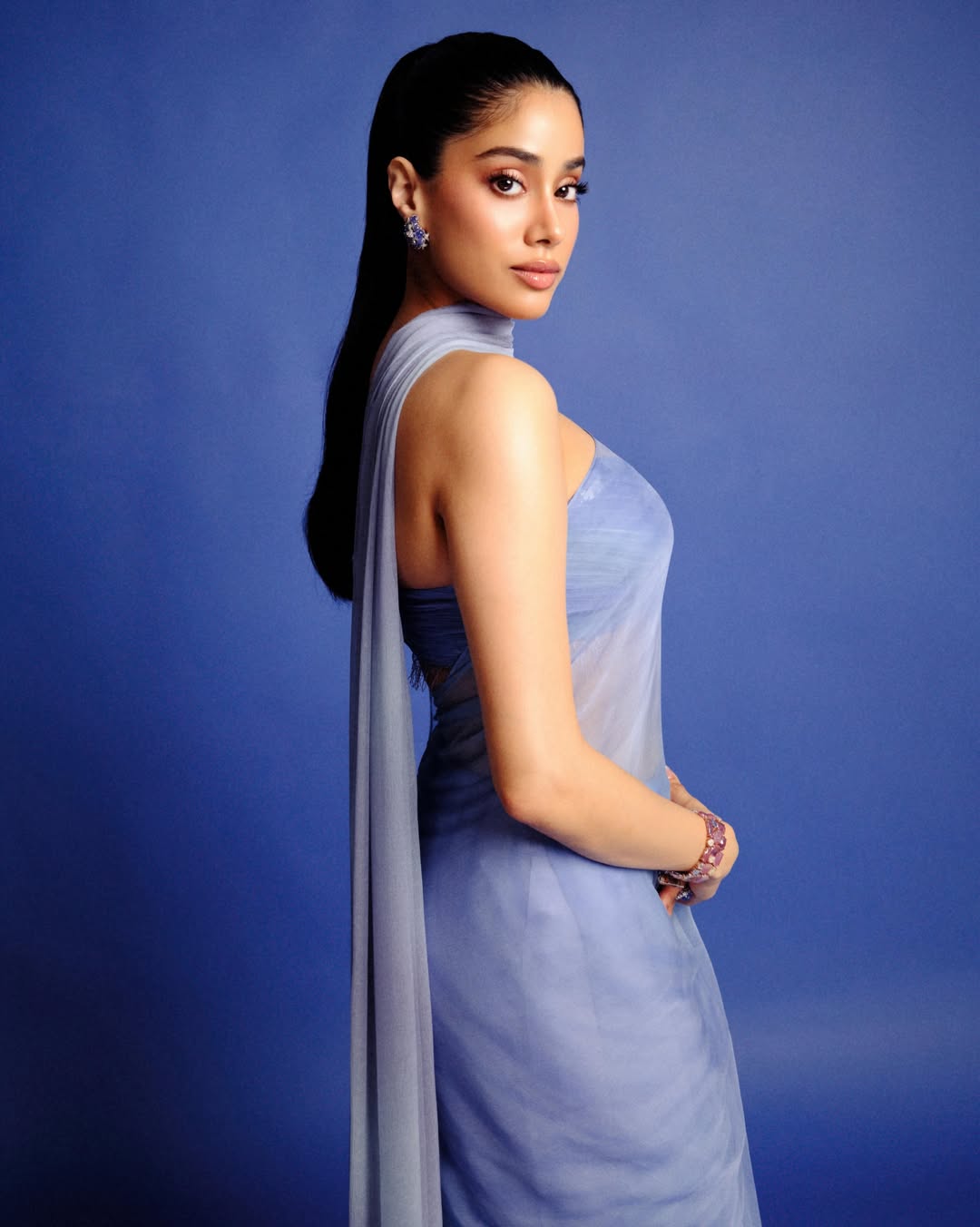 Janhvi Kapoor poses in a flowing, sheer light blue garment against a vibrant blue background, showcasing elegant draping.