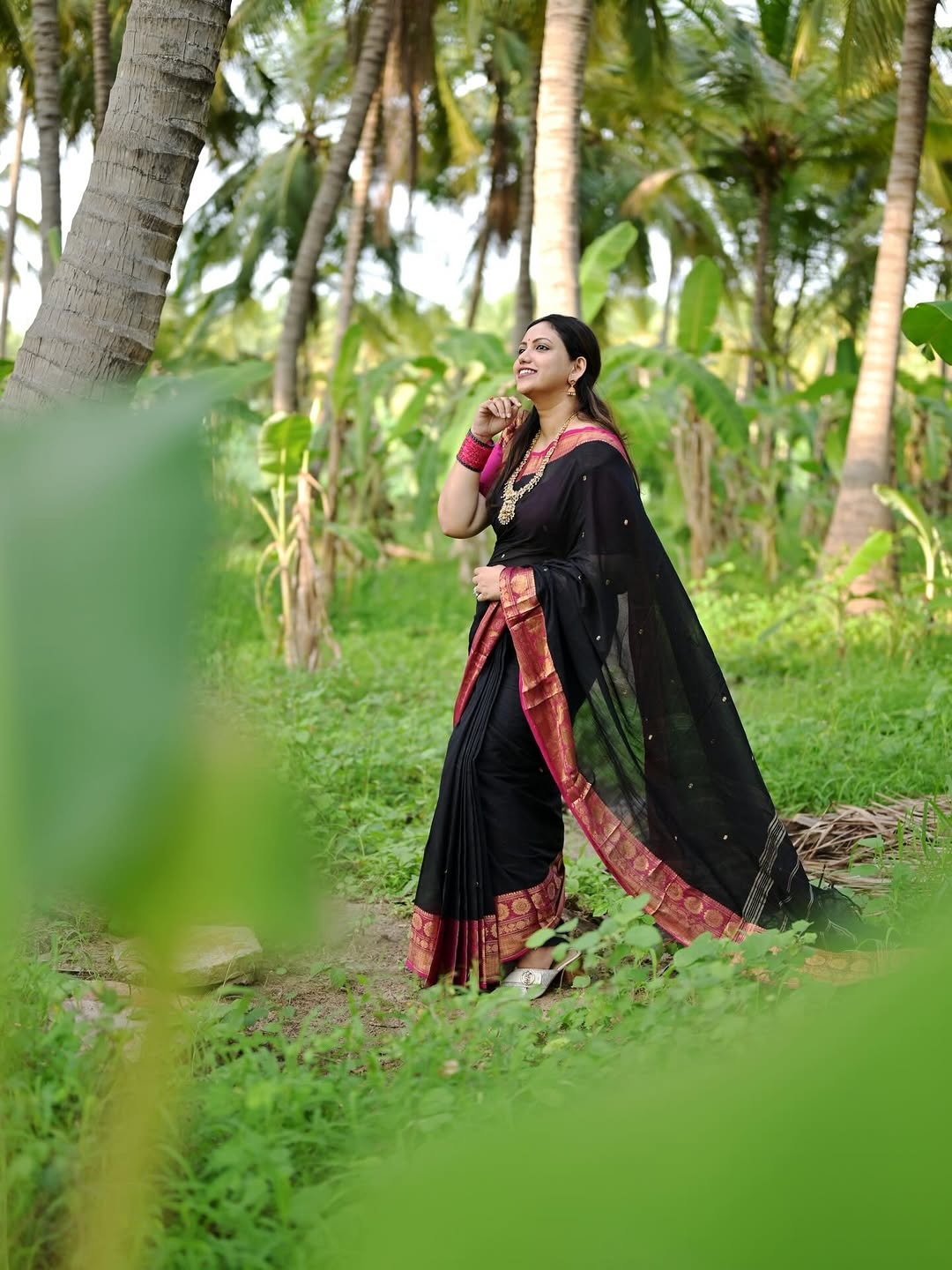 Joy Crizilda standing outdoors smiling in a black saree with a magenta border and gold jewelry.