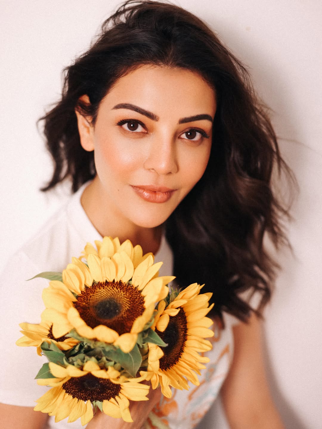 Kajal Aggarwal sitting with a bouquet of yellow sunflowers against a light background, wearing a white crop top and a light blue polka dot skirt.