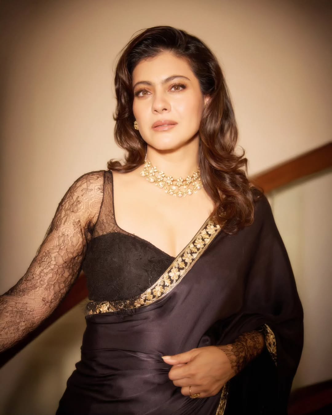 Kajol in a close-up portrait with long, dark, wavy hair and embellished attire, looking directly at the camera with a warm expression.