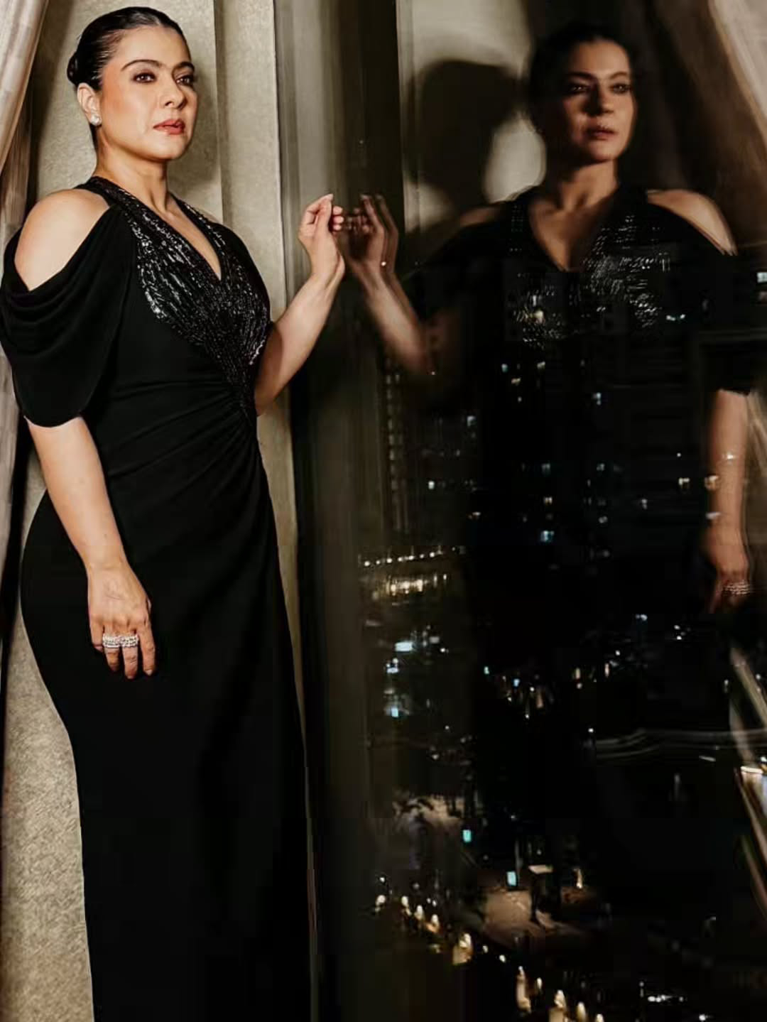 Kajol stands in a full-length portrait wearing a long, flowing black gown with sparkling embellishments, leaning against a warm wooden background.