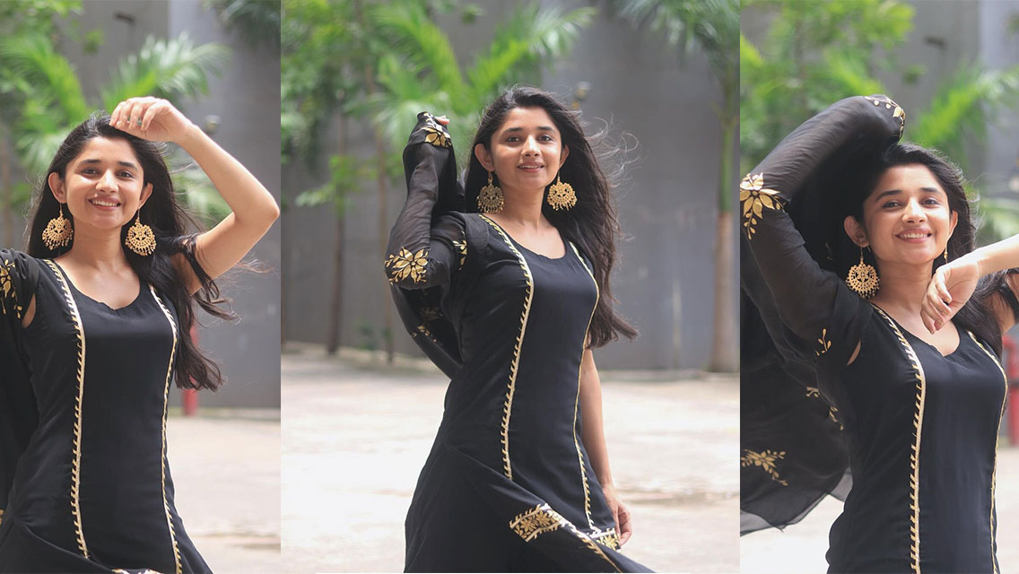 Kanika Mann stands outdoors wearing a flowing black garment with gold detailing and draped fabric, posing in bright daylight.