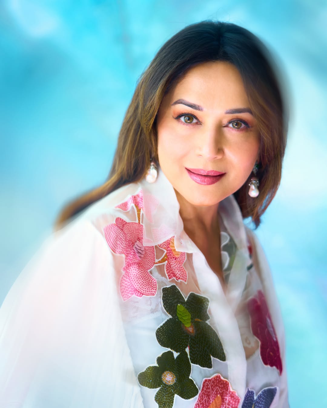 Madhuri Dixit stands in a flowing outfit against soft, draped fabric, showcasing a vibrant floral print and relaxed pose.