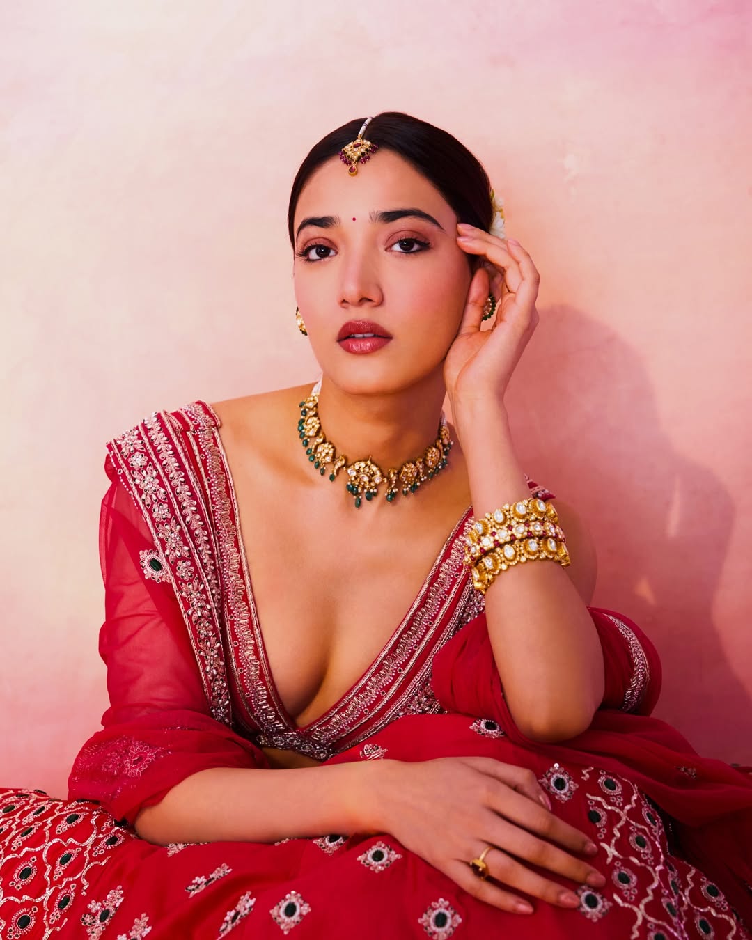 Medha Shankr posing in a red lehenga against a soft pink background, smiling softly.