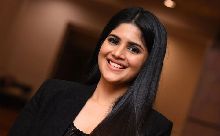 Megha Akash in a full-length portrait wearing an all-black ensemble and standing against a light background.
