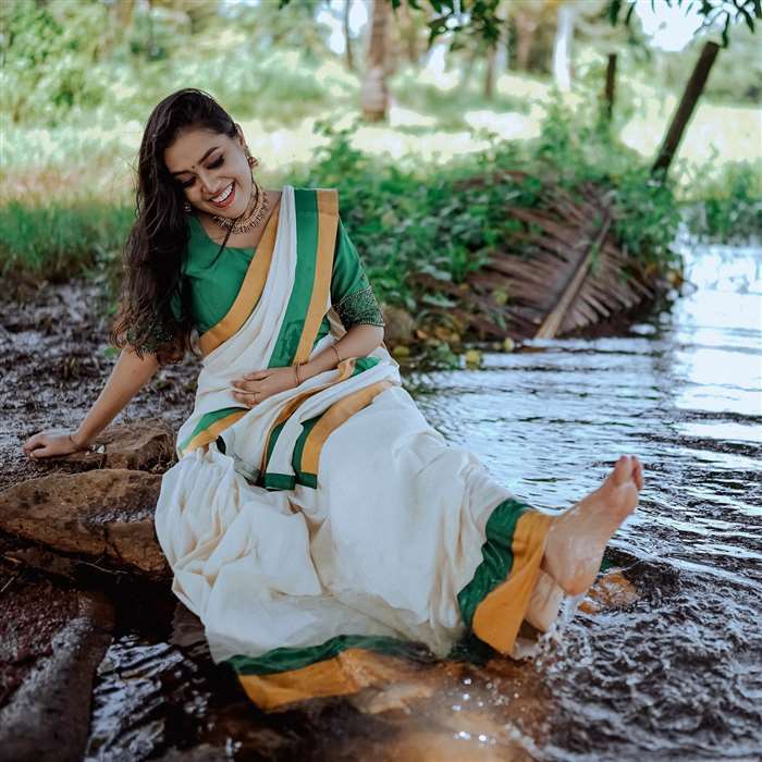 Mithu Vigil with long, reddish-brown hair, wearing a flowing green saree with gold accents, leaning against a vertical structure outdoors.