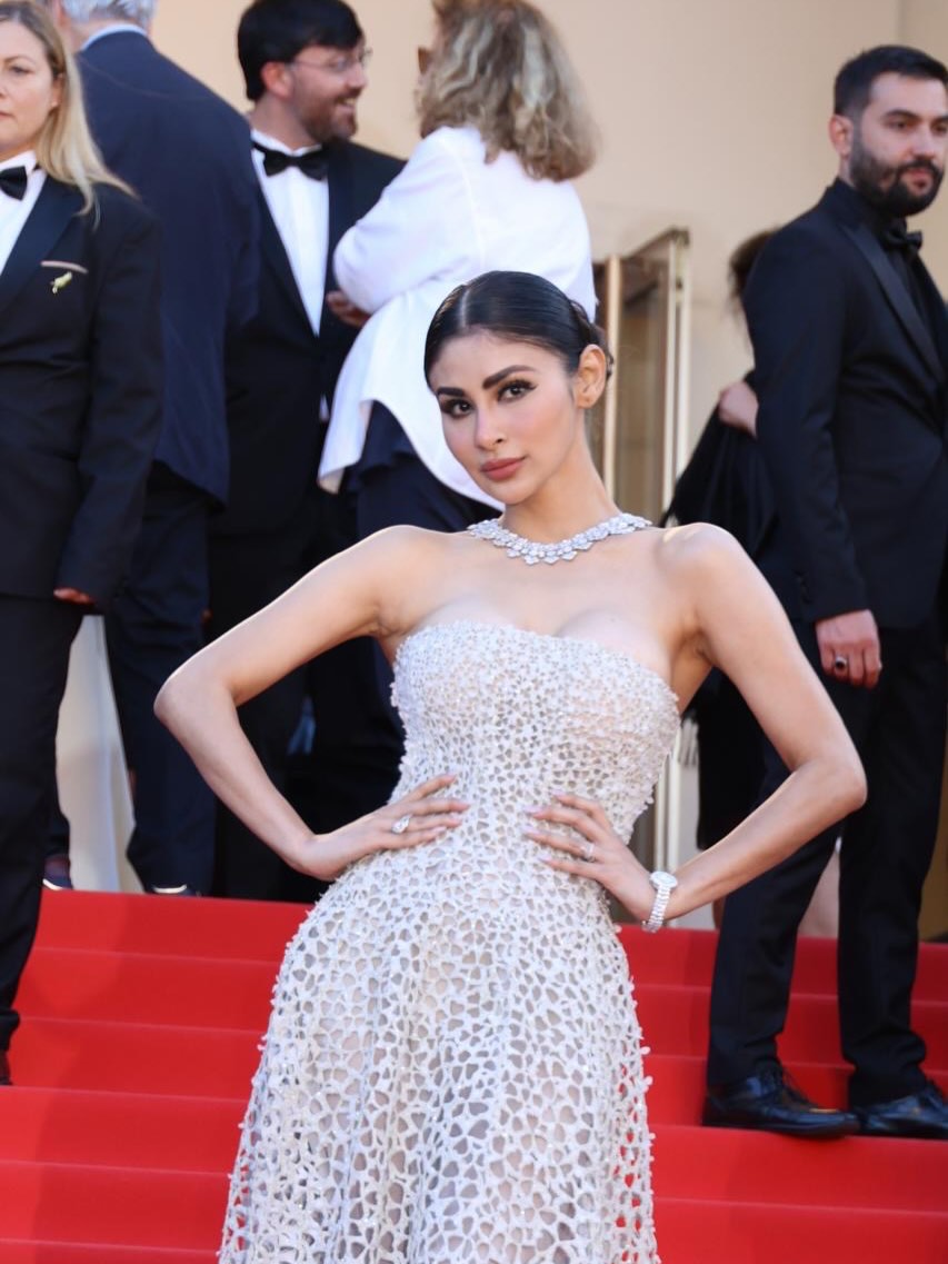 Mouni Roy seated inside a car wearing a light-colored, intricately textured lace gown, looking at the camera.
