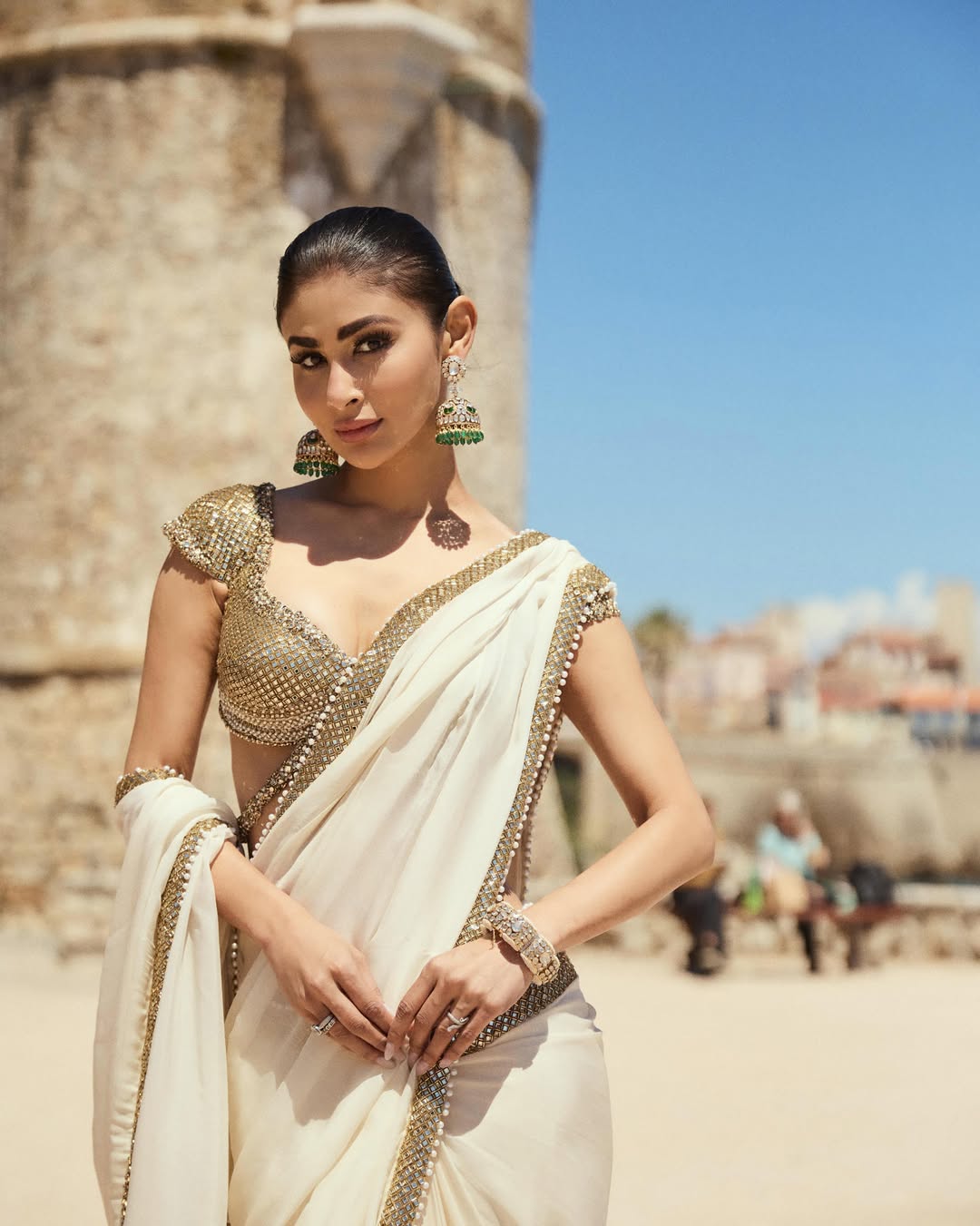 Mouni Roy models an exquisite ivory and gold saree, radiating grace against a serene beach backdrop.