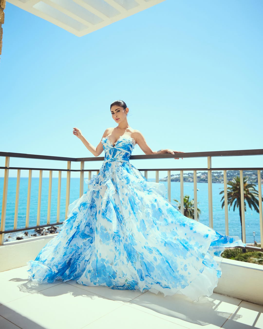 Mouni Roy standing outdoors leaning against a railing overlooking the bright blue ocean and clear sky.