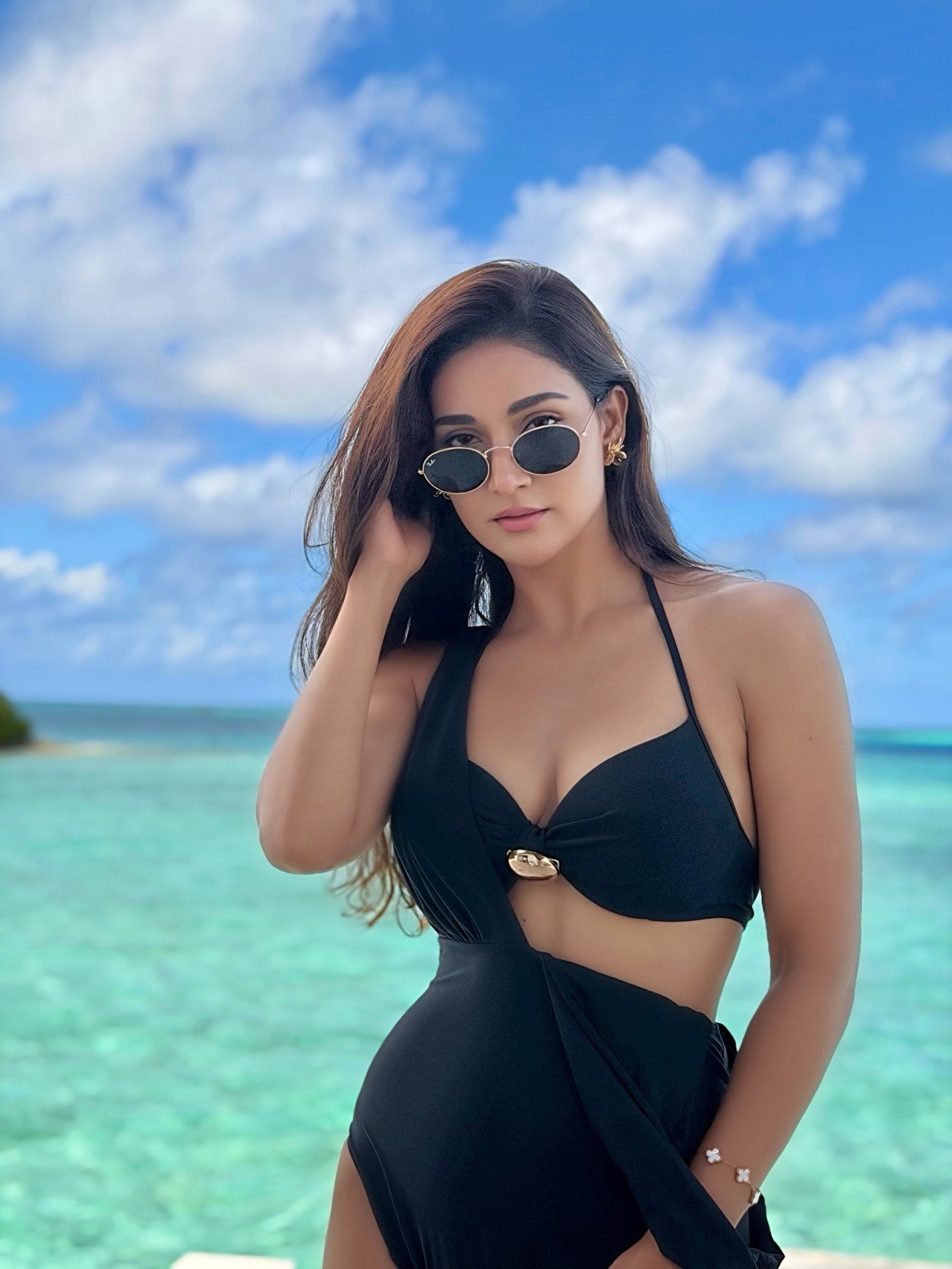 Mukti Mohan stands on a wooden pier over clear turquoise water against a backdrop of blue sky and clouds. - Photos & Updates