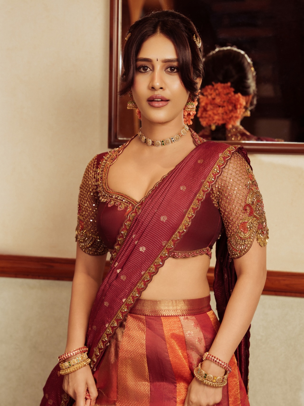 Nabha Natesh is captured looking into an ornate mirror while wearing traditional maroon and gold ethnic attire.