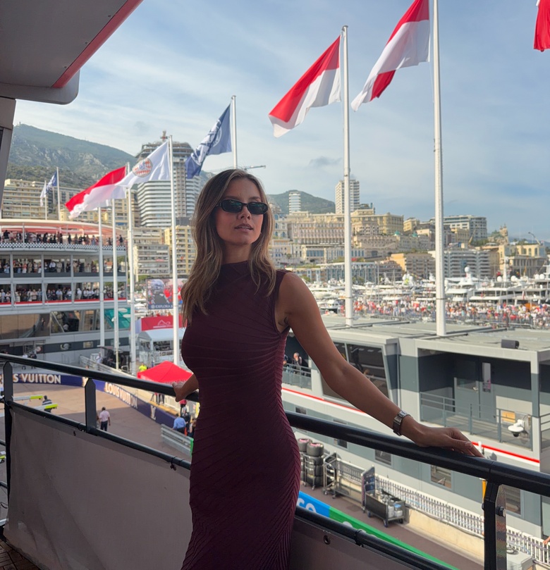 Nicola Cavanis standing indoors in a flowing beige dress overlooking a busy harbor and city landscape.