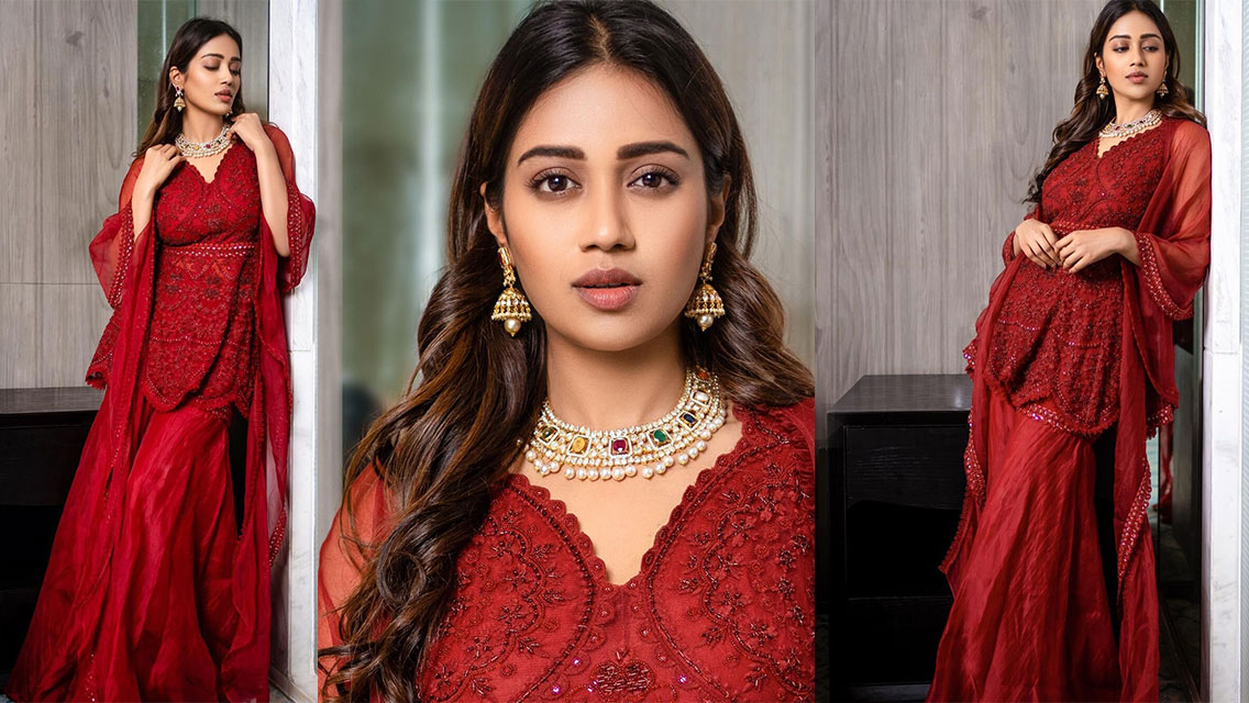 Nivetha Pethuraj is wearing a flowing, deep red or maroon garment with intricate embroidery, leaning against a light-colored surface indoors.
