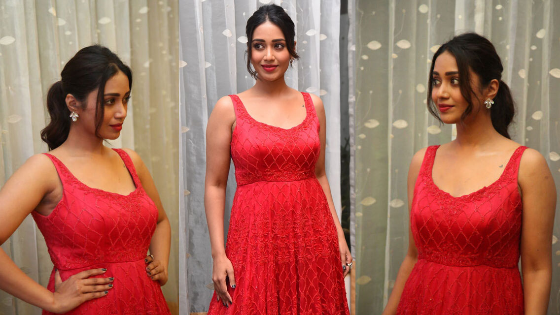 Nivetha Pethuraj smiles warmly in a vibrant red, heavily embellished outfit against a light fabric backdrop.