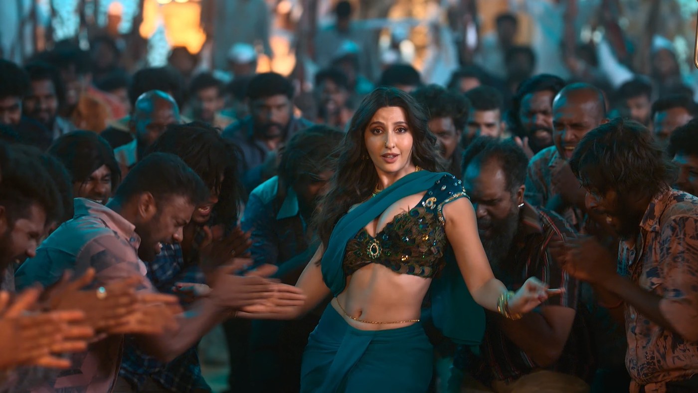 Nora Fatehi performing on a brightly lit stage wearing a sparkling pink and magenta outfit and holding a microphone.