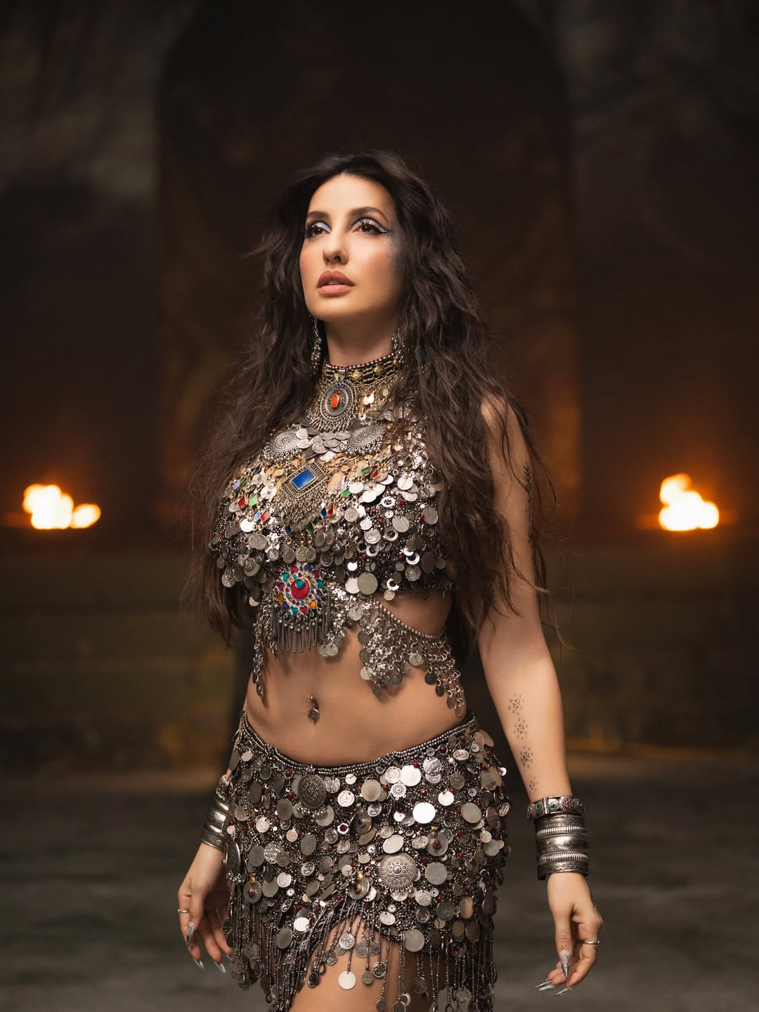 Nora Fatehi in an ornate, heavily embellished garment with gold-toned jewelry and metallic bangles.