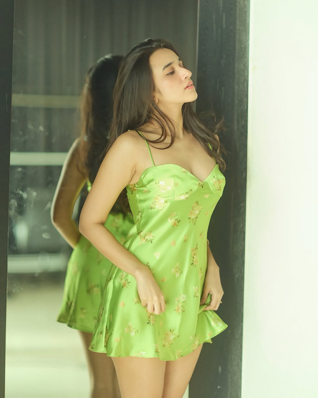 Nuveksha is captured leaning against a dark vertical surface in a light green dress with floral print and gold accessories.