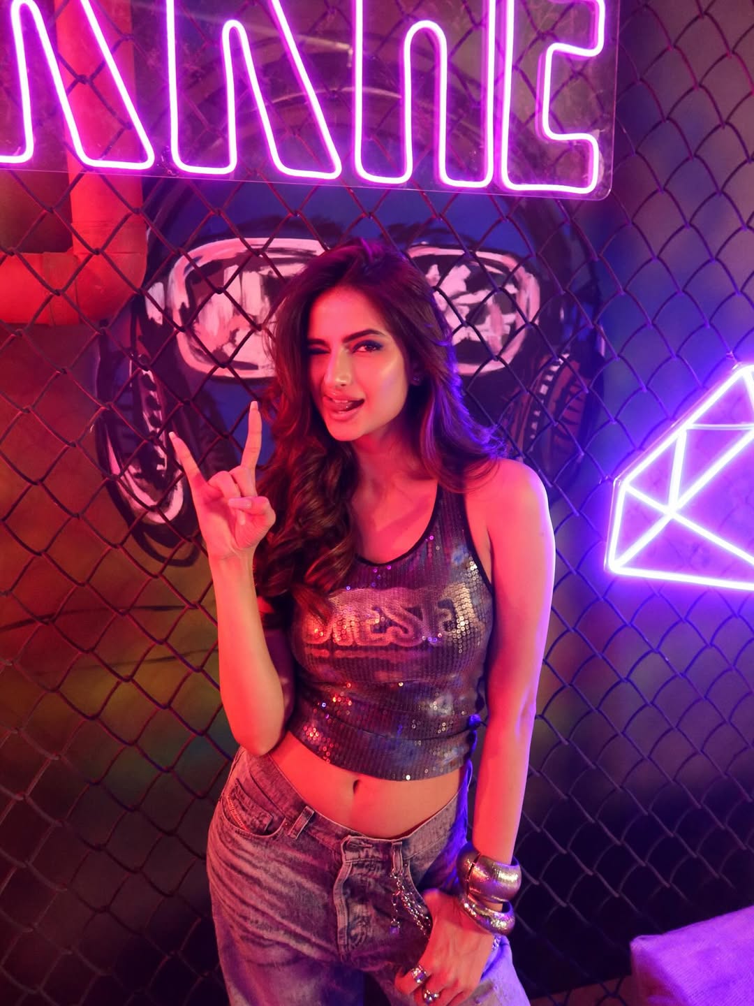 Palak Tiwari stands in front of a colorful mural wearing a shimmering, iridescent sleeveless top and light blue denim jeans.