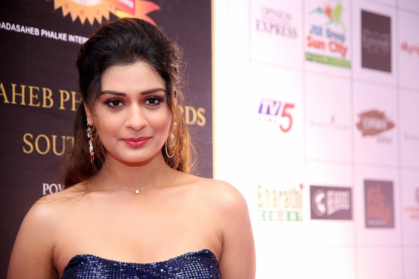Payal Rajput posing indoors wearing a shimmering navy blue, heavily embellished strapless garment and gold hoop earrings.