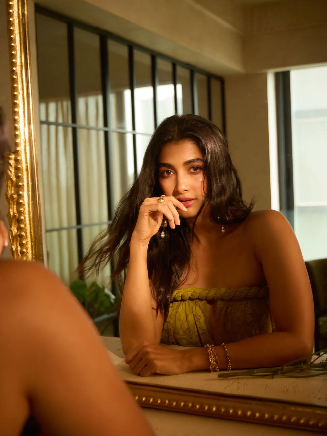 Pooja Hegde seated indoors wearing a flowing garment in golden yellow-green with intricate patterns.