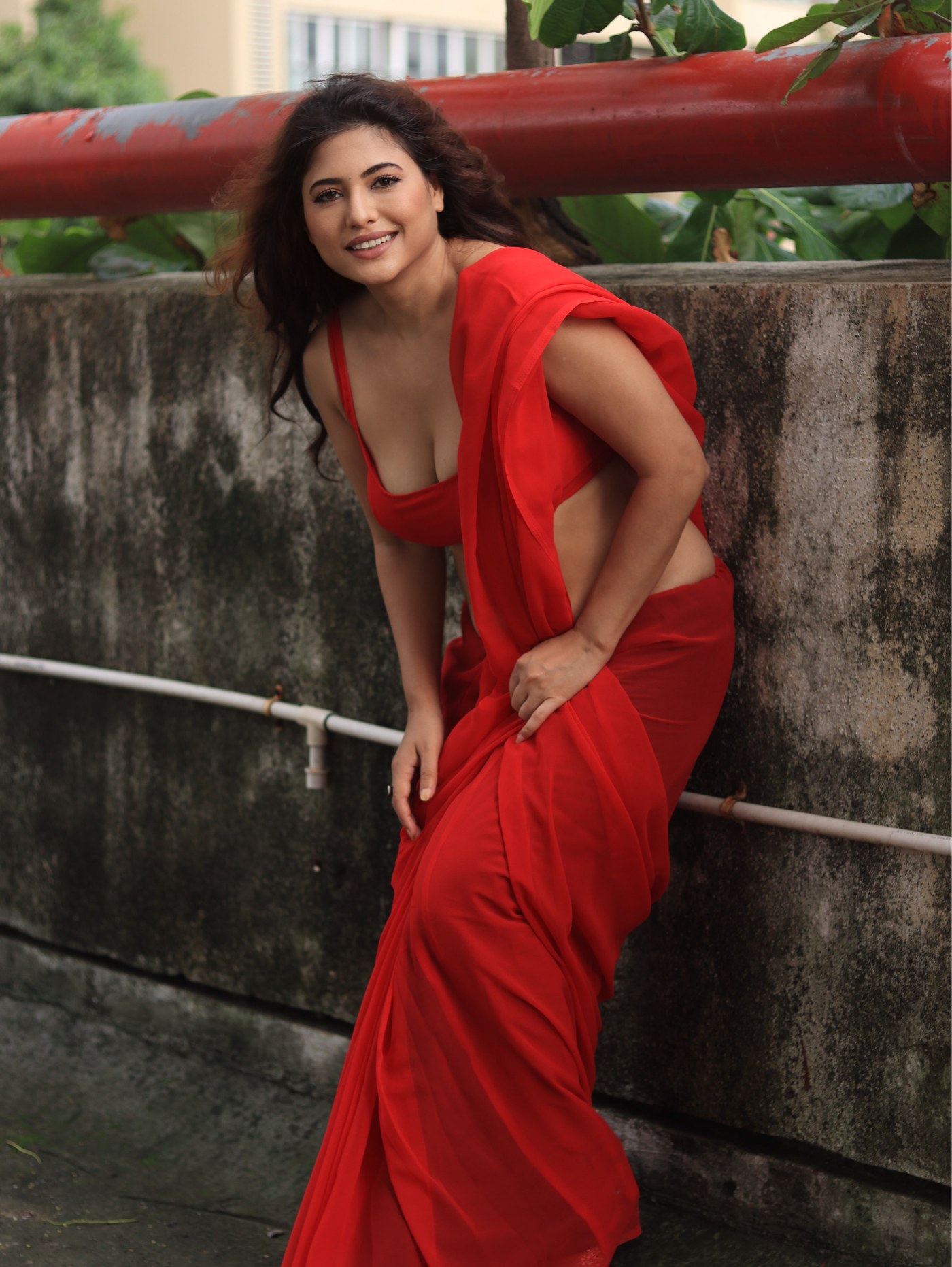 Poonam Rajput stands in a striking red ensemble against weathered architectural pillars.