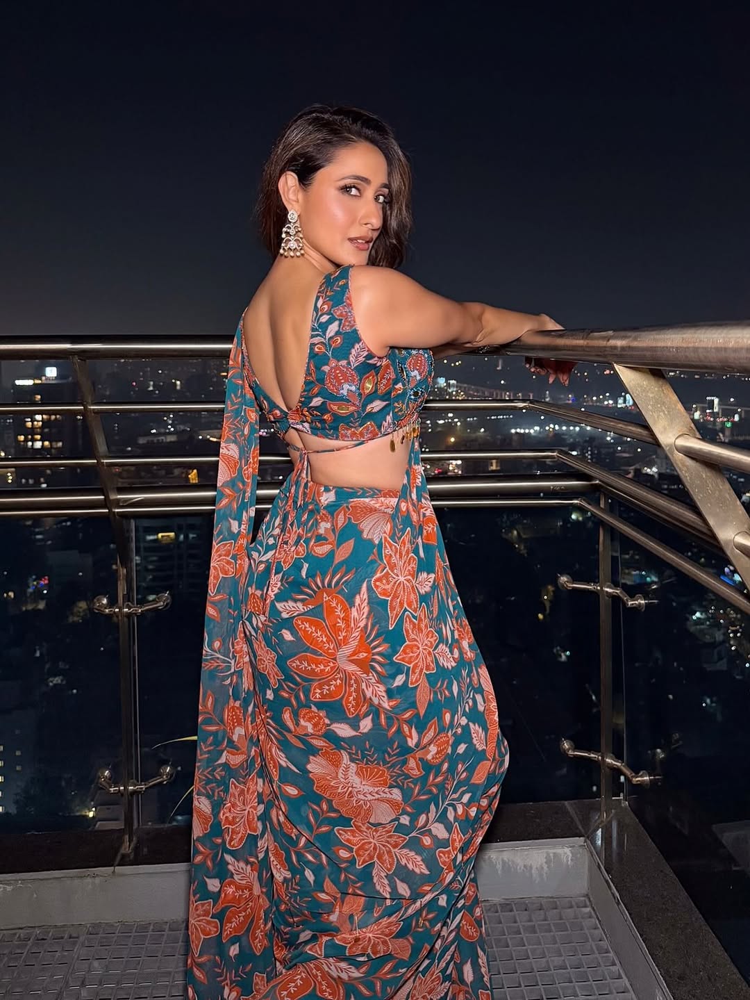 Pragya Jaiswal posing against a textured, warm-toned background wearing a teal and orange patterned garment with layered necklaces.