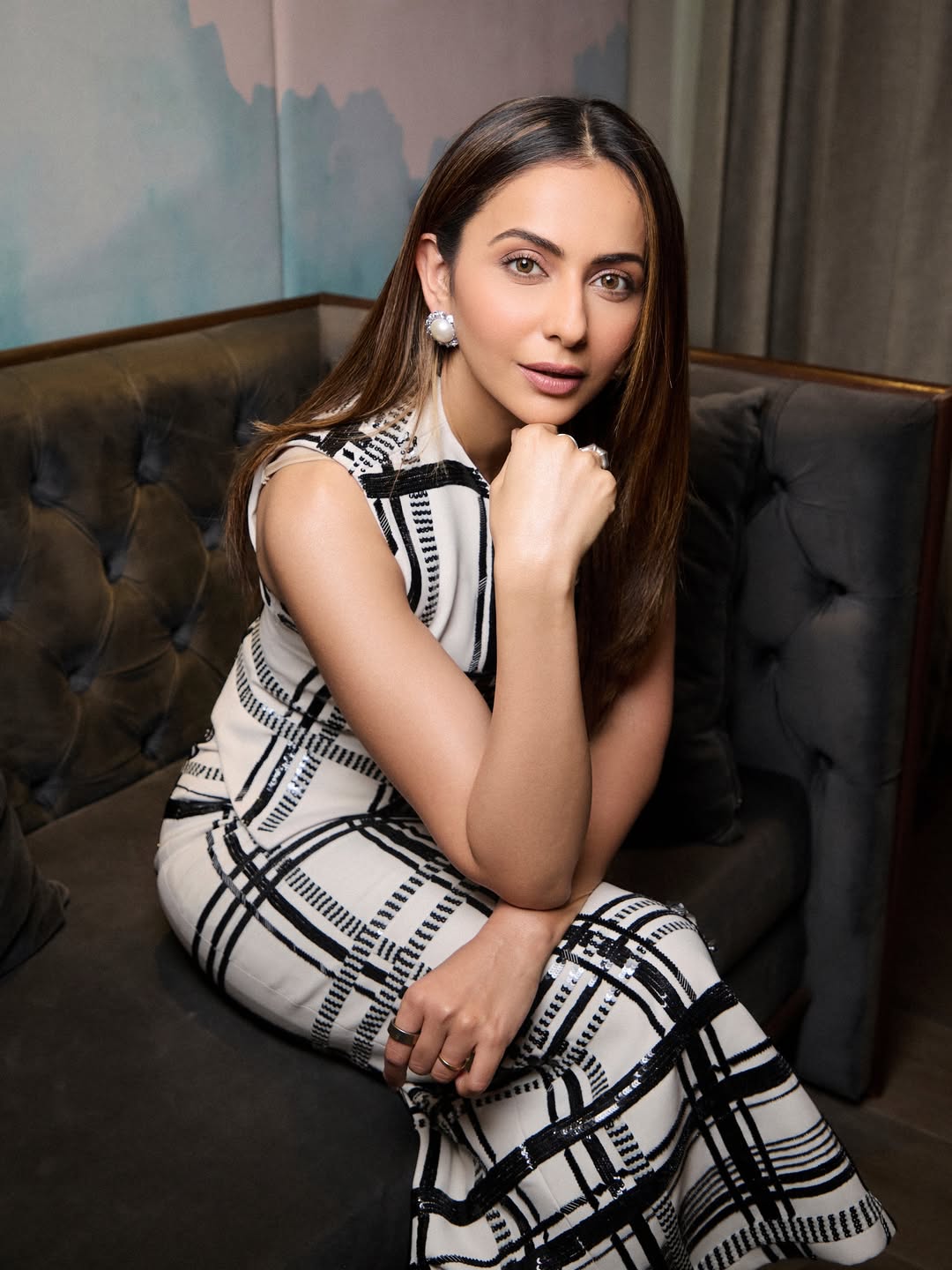 Rakul Preet Singh poses indoors next to a tufted sofa wearing a fitted black and white patterned dress and black heels.