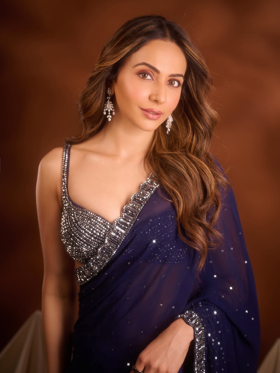 Rakul Preet Singh is featured in a portrait wearing an embellished blue saree with flowing waves and silver jewelry.
