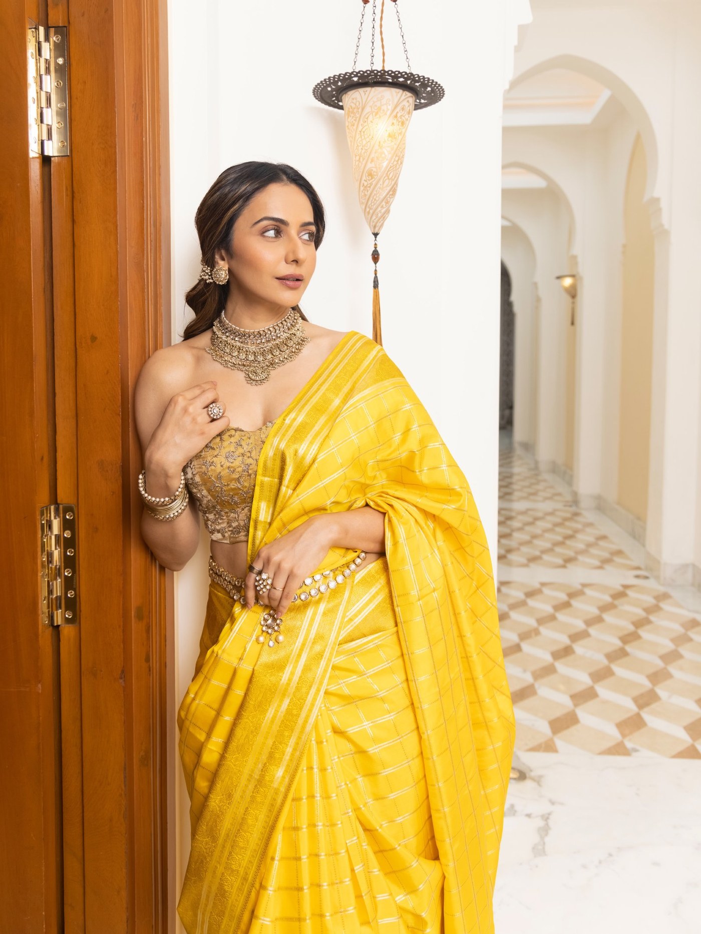 Rakul Preet Singh poses gracefully in a vibrant mustard yellow saree and elaborate gold-toned jewelry on an ornate staircase.