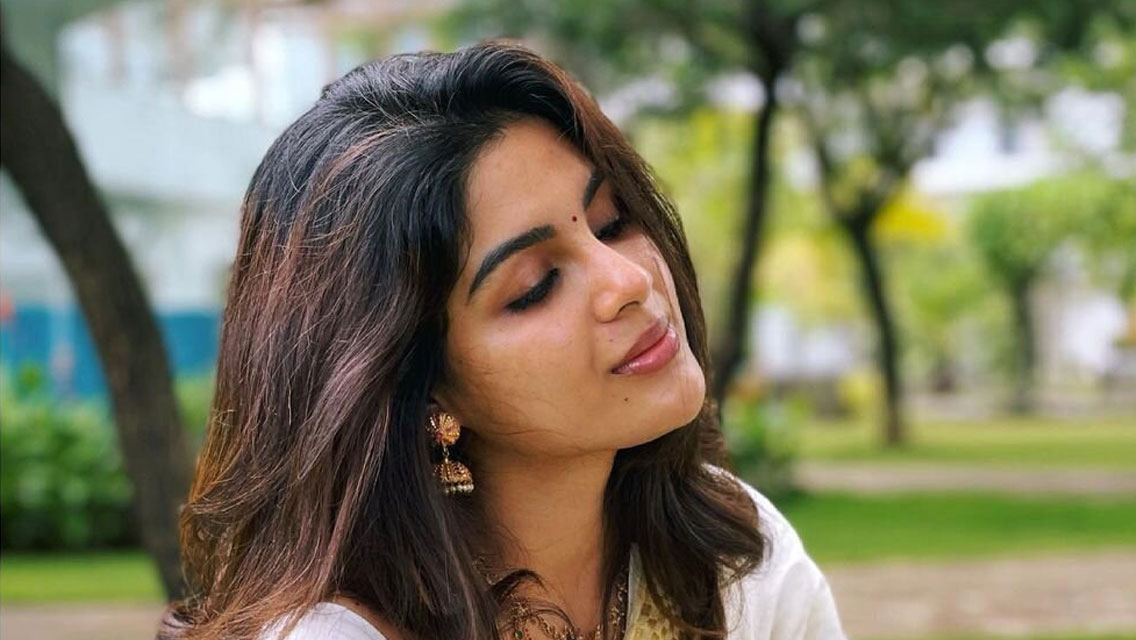 Samyuktha Menon lying on bright green grass wearing a cream saree with a golden border in a garden setting.