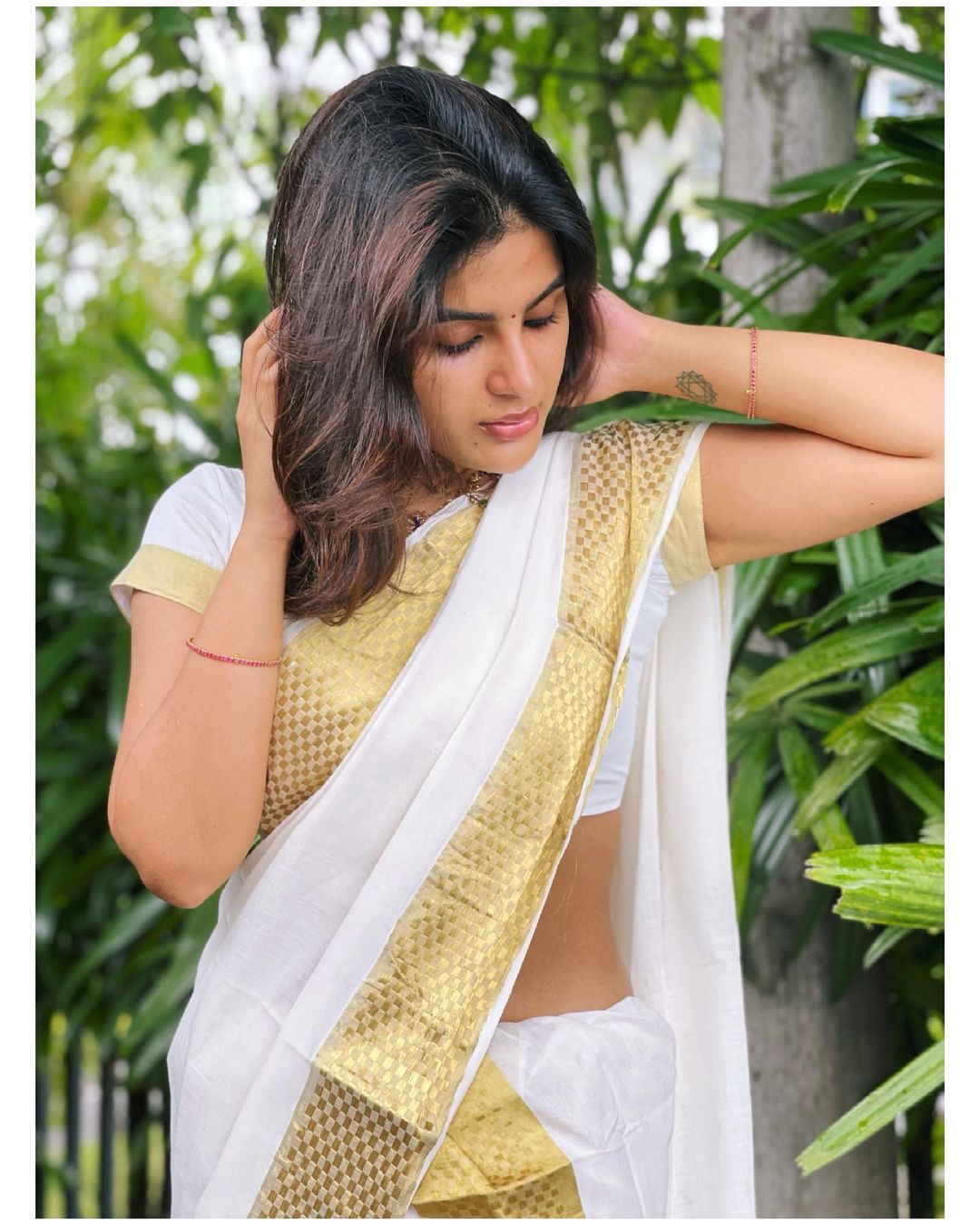 Samyuktha Menon lying on bright green grass wearing a cream saree with a golden border in a garden setting.